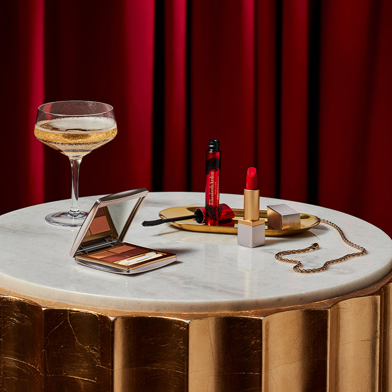 Makeup items including a compact mirror, lipsticks, and a glass on a marble table with a red curtain background.
