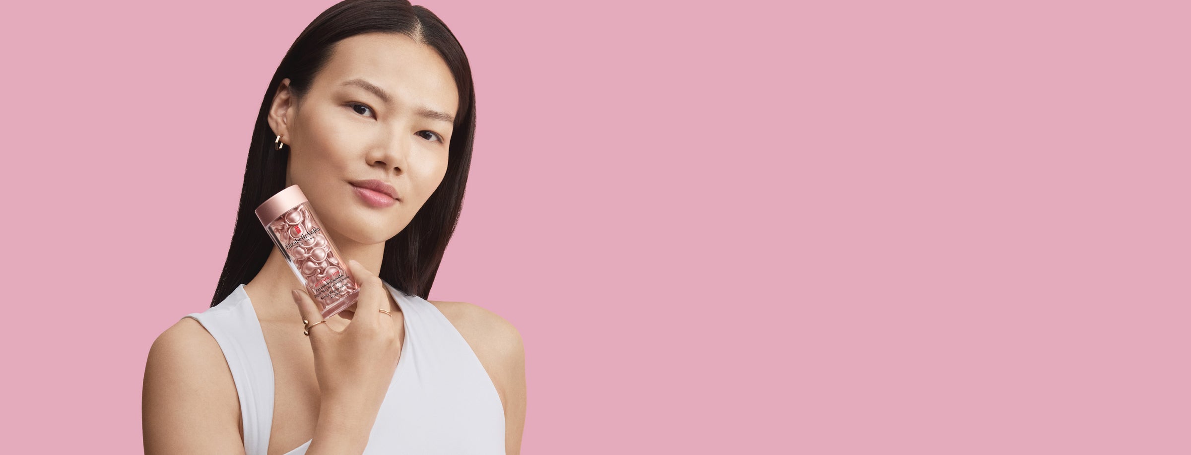 Woman holding a pink skincare product container containing capsules against a pink background