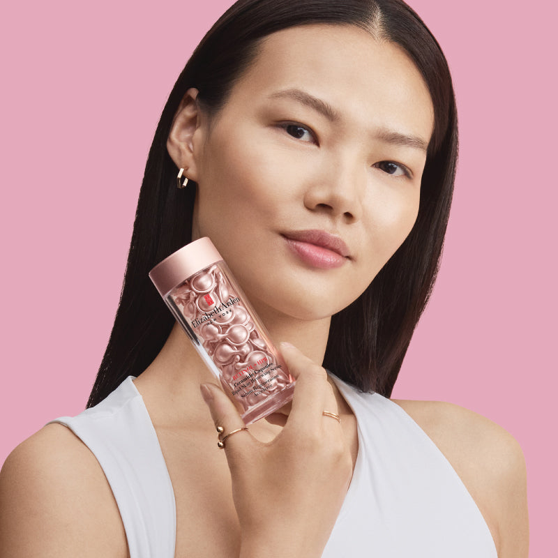 Woman holding a pink skincare product container containing capsules against a pink background