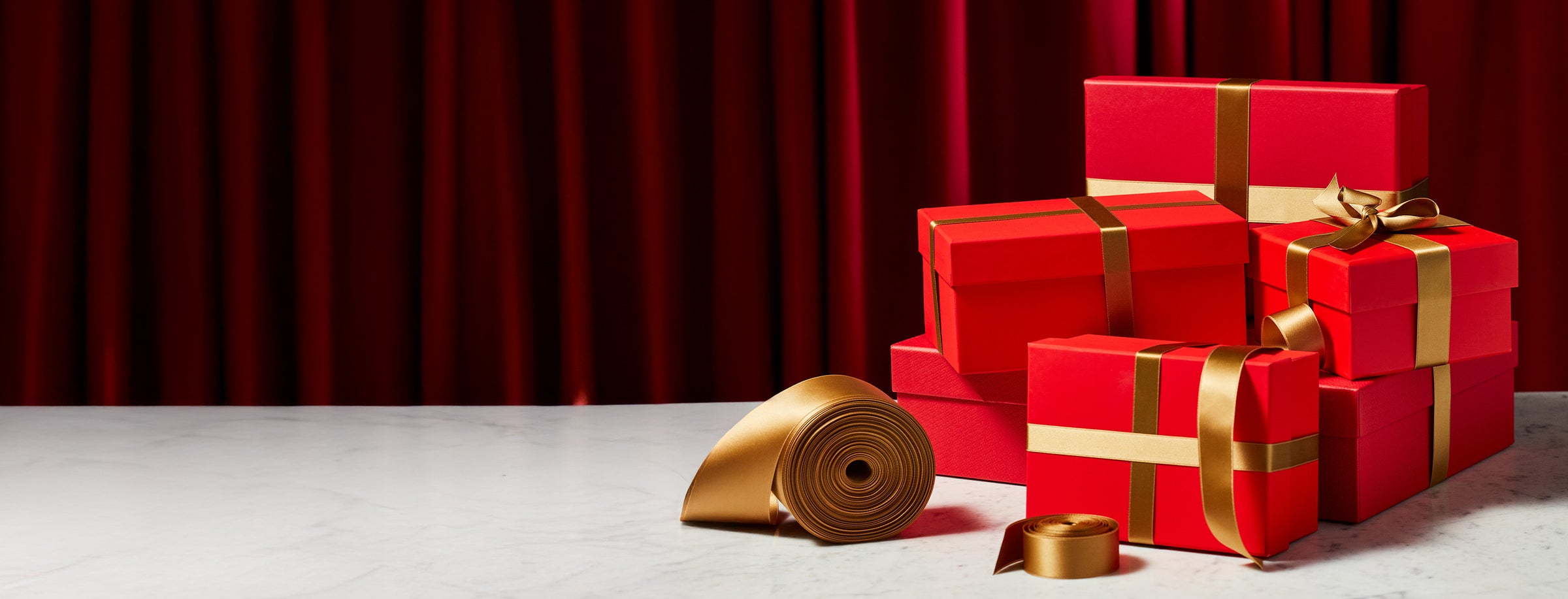 Stack of red gift boxes with gold ribbons on a white surface with a red curtain background