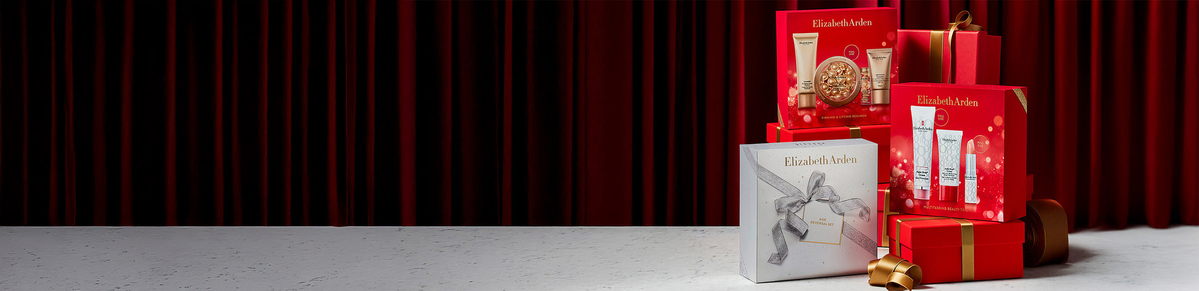 Elizabeth Arden gift sets featured on red gift boxes with gold ribbon on a red curtain background