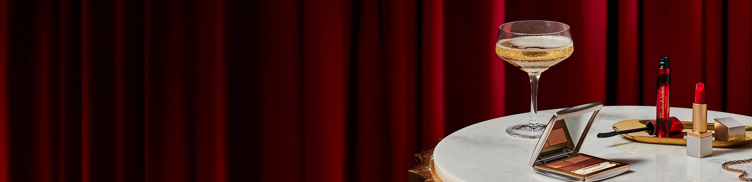 Makeup items including a lipstick, and mascara on a marble table with a red curtain background.