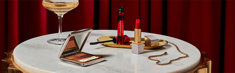 Makeup items including a lipstick, and mascara on a marble table with a red curtain background.