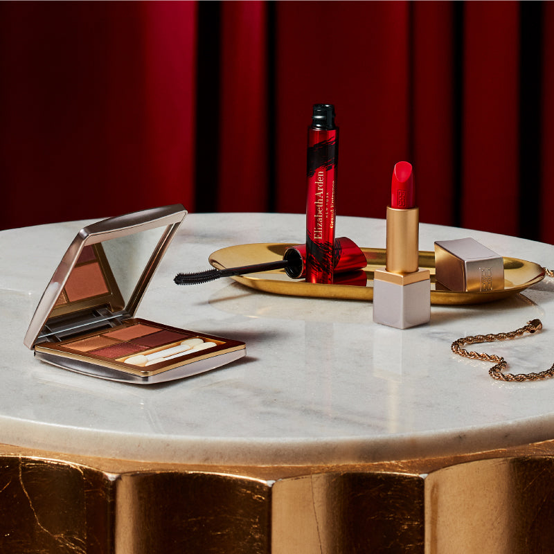 Makeup products including a compact, lipstick, and mascara on a marble table with a red curtain background.