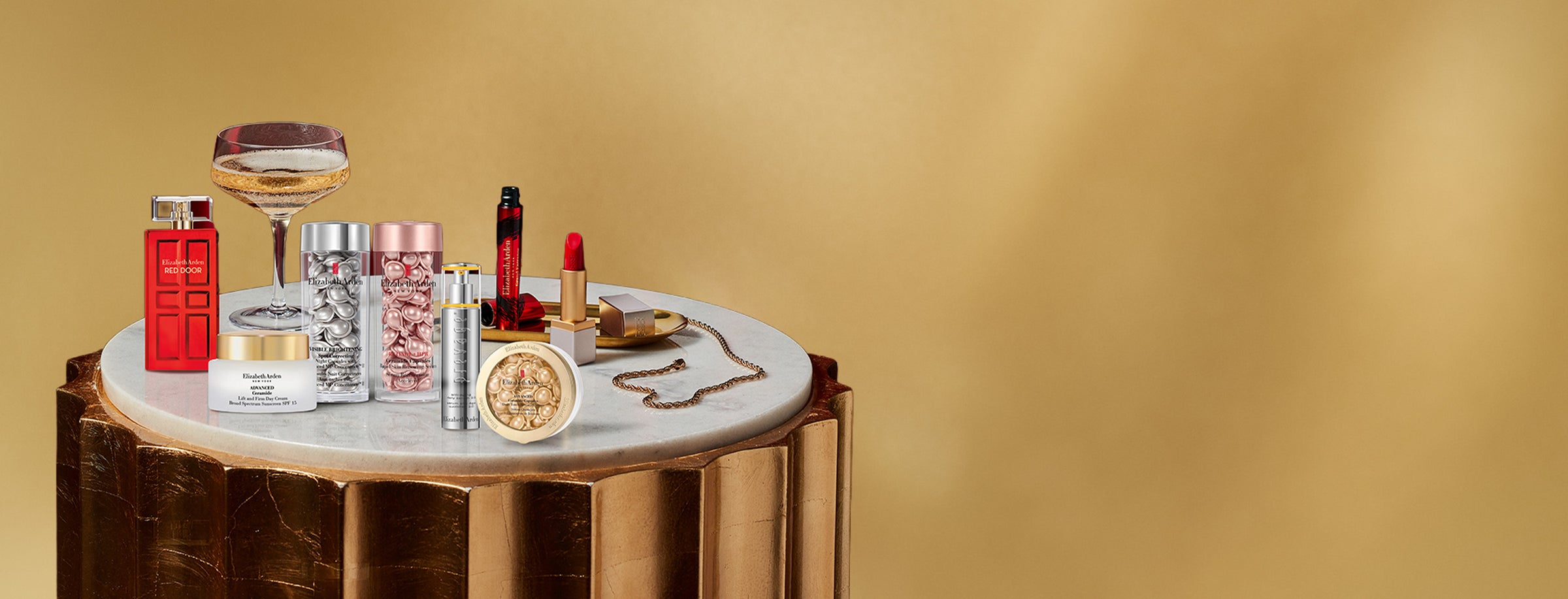 Tabletop display with beauty products, jewelry, and a glass of champagne on a gold background