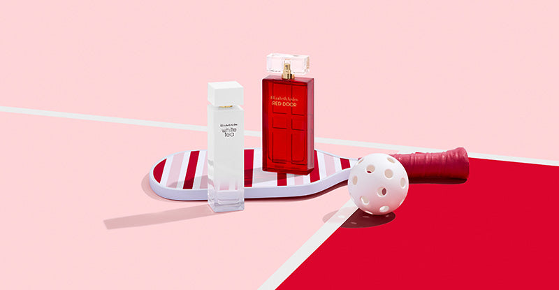 Two perfume bottles on stripe pickleball paddle on a pink and red background next to a ball