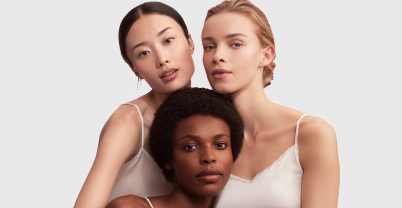Three women of different ethnicities wearing white tank tops against a light gray background