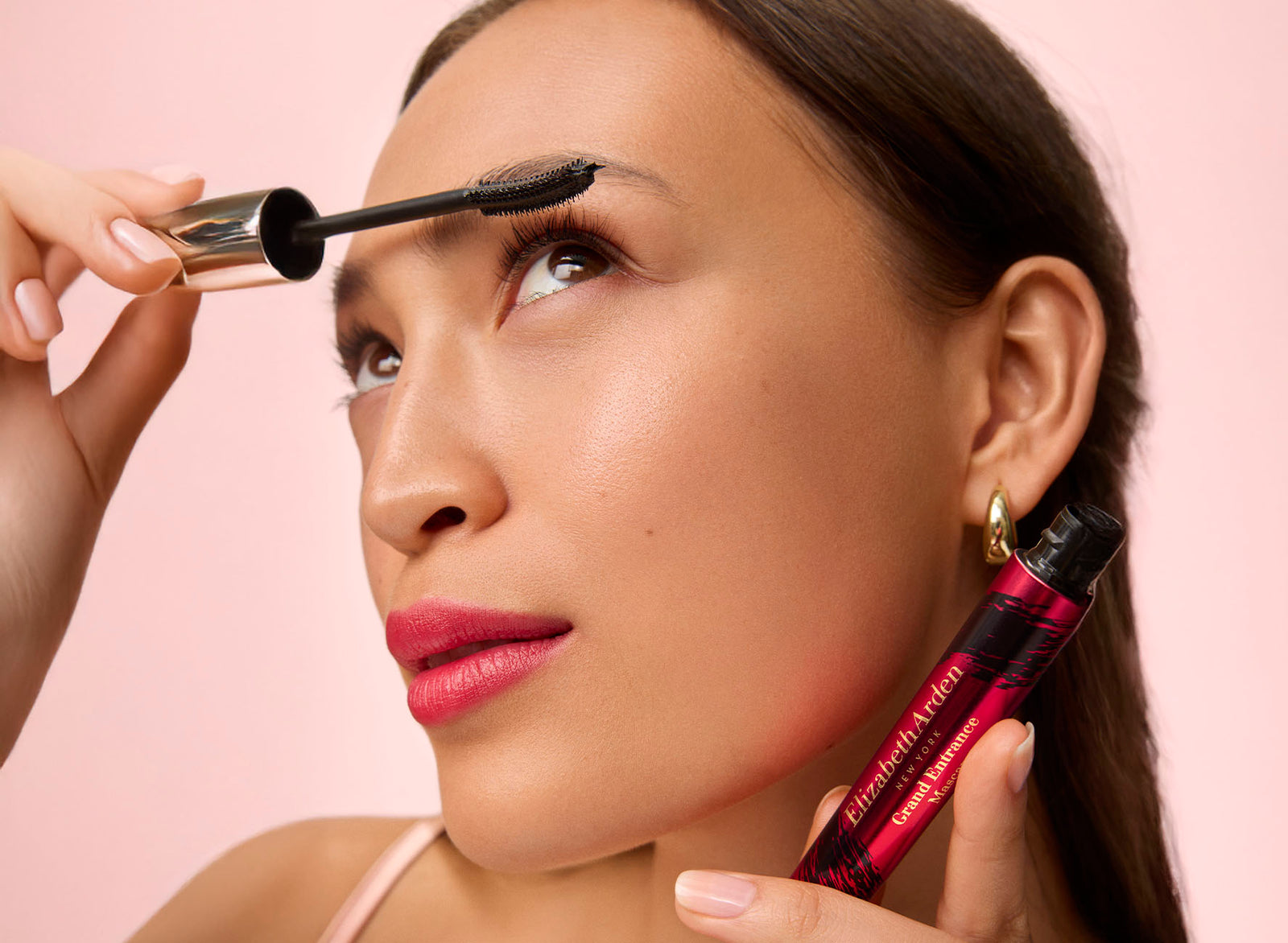 Woman applying makeup with a brush and mascara on a pink background