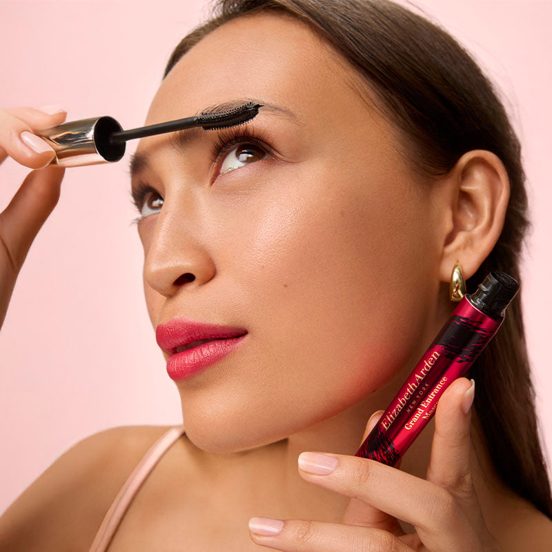 Woman applying makeup with a brush and mascara on a pink background