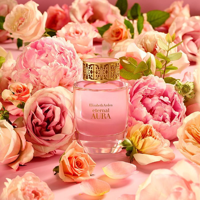 Pink Elizabeth Arden Eternal Aura perfume bottle surrounded by pink and peach roses on a pink background
