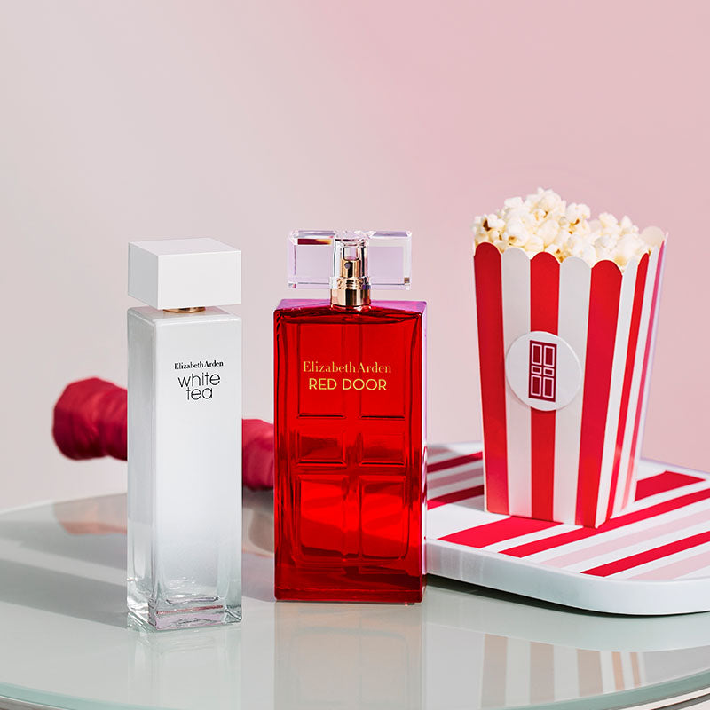 Two perfume bottles and a popcorn container on a glass surface with a pink background