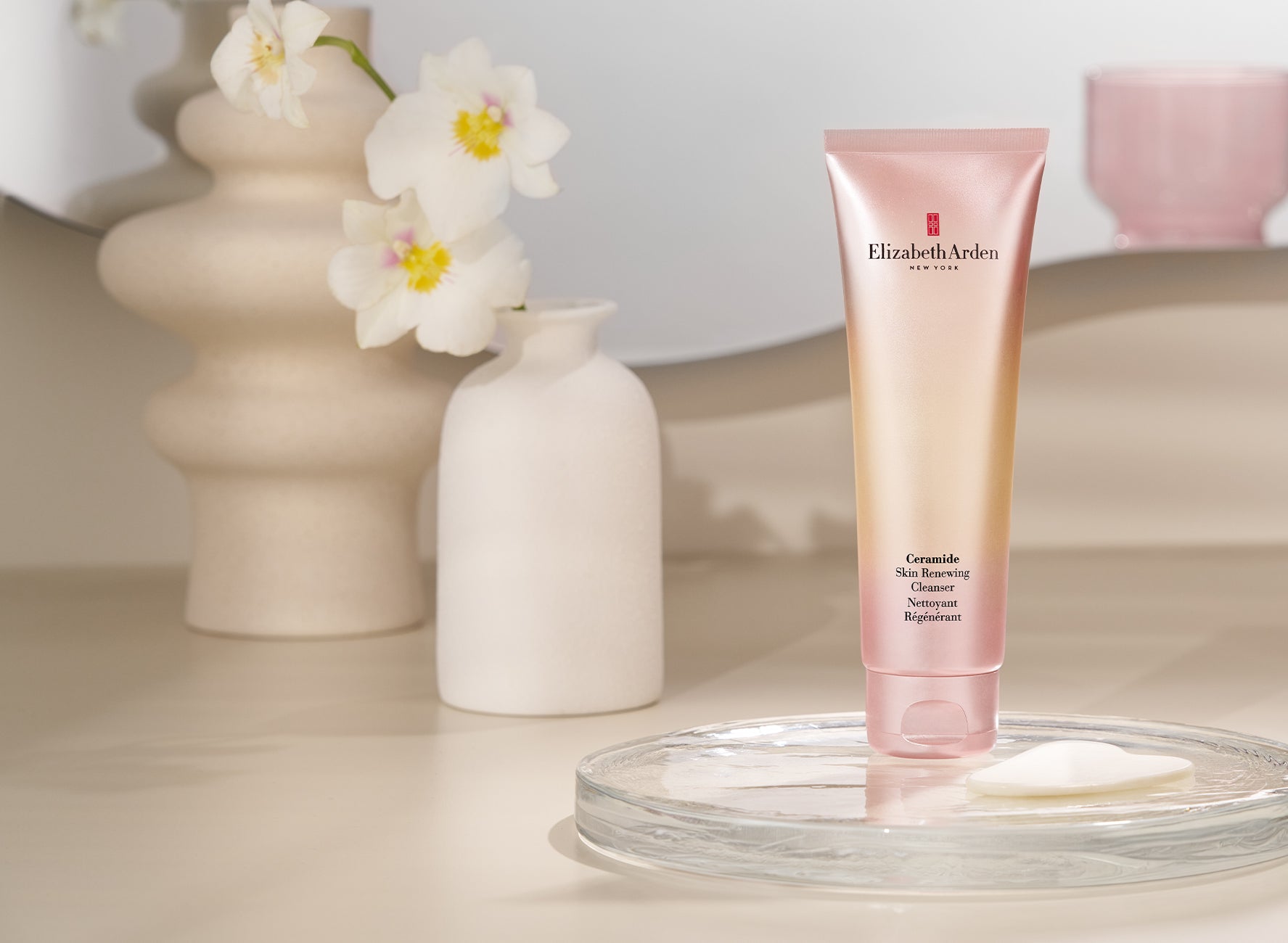 Elizabeth Arden Ceramide Cleanser on a clear tray against a beige background next to 2 vase with flowers.