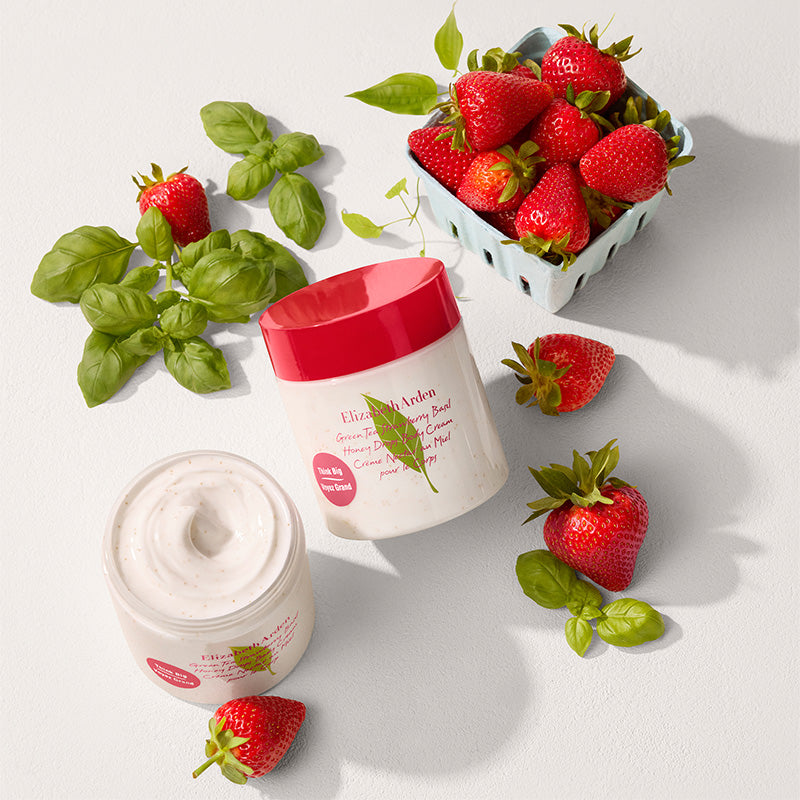 Two jars of cream with a red lid, surrounded by strawberries and basil leaves on a light background.