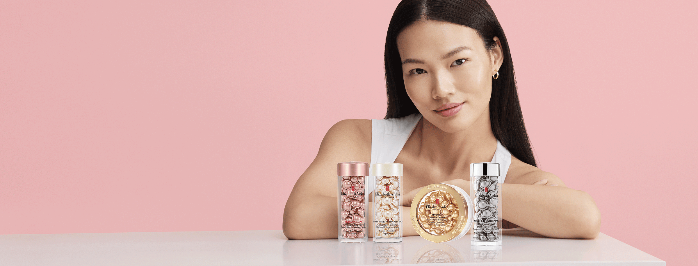 Woman sitting at a table with Elizabeth Arden capsules serum products against a pink background