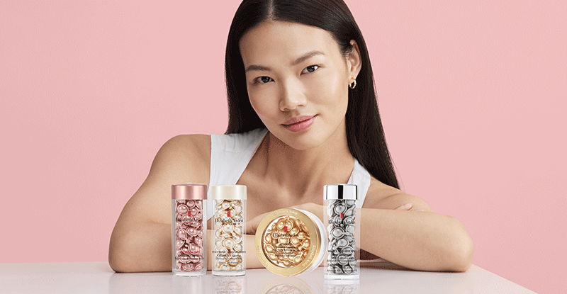 Woman sitting at a table with Elizabeth Arden capsules serum products against a pink background