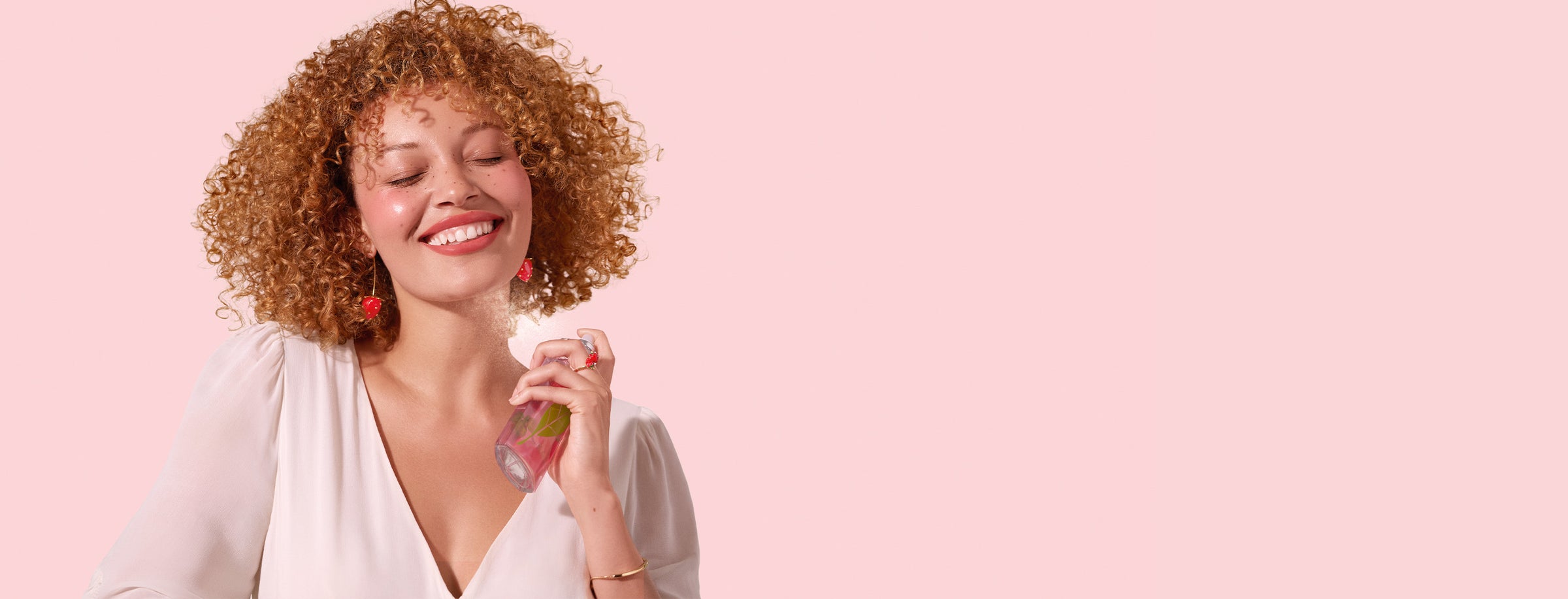 Model with curly red hair, smiling while spray perfume on her neck