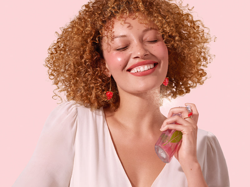 Model with curly red hair, smiling while spray perfume on her neck