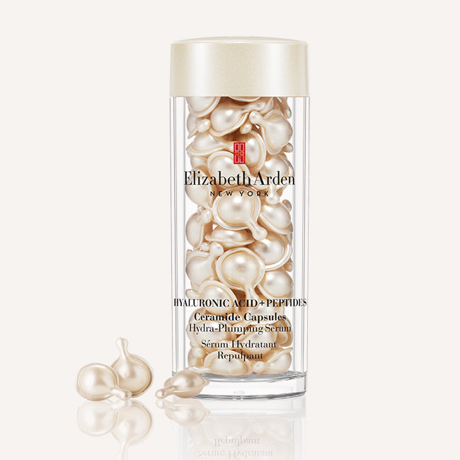 Elizabeth Arden Hyaluronic Acid + Peptides Ceramide Capsules in a clear cylindrical bottle with a cream-colored cap, shown on a light beige background with a few capsules resting outside the bottle.