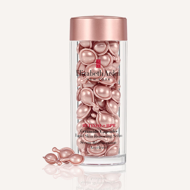 Elizabeth Arden Retinol + HPR Ceramide Capsules in a clear cylindrical bottle with a metallic rose gold cap, with three capsules resting outside on a reflective surface against a light beige background.