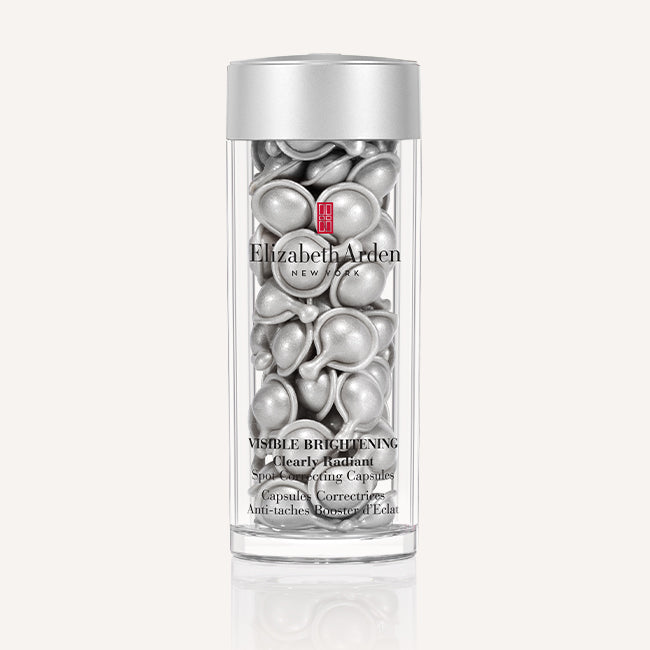 A clear cylindrical bottle of Elizabeth Arden Visible Brightening Corrective Capsules with a metallic silver cap, showing the silver capsule contents inside, on a light beige background.