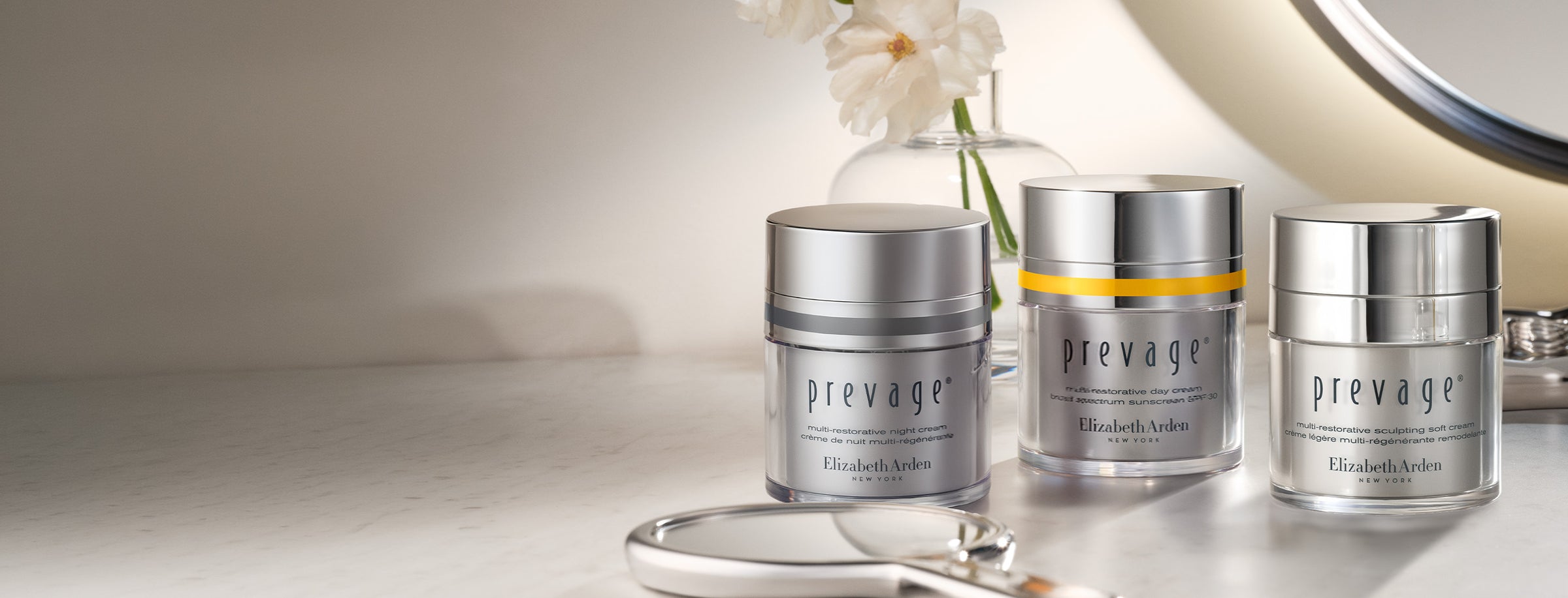 Three Elizabeth Arden Prevage Multi-restorative moisturizers are arranged on a reflective surface with a vase of white flowers and part of a round mirror in the background.