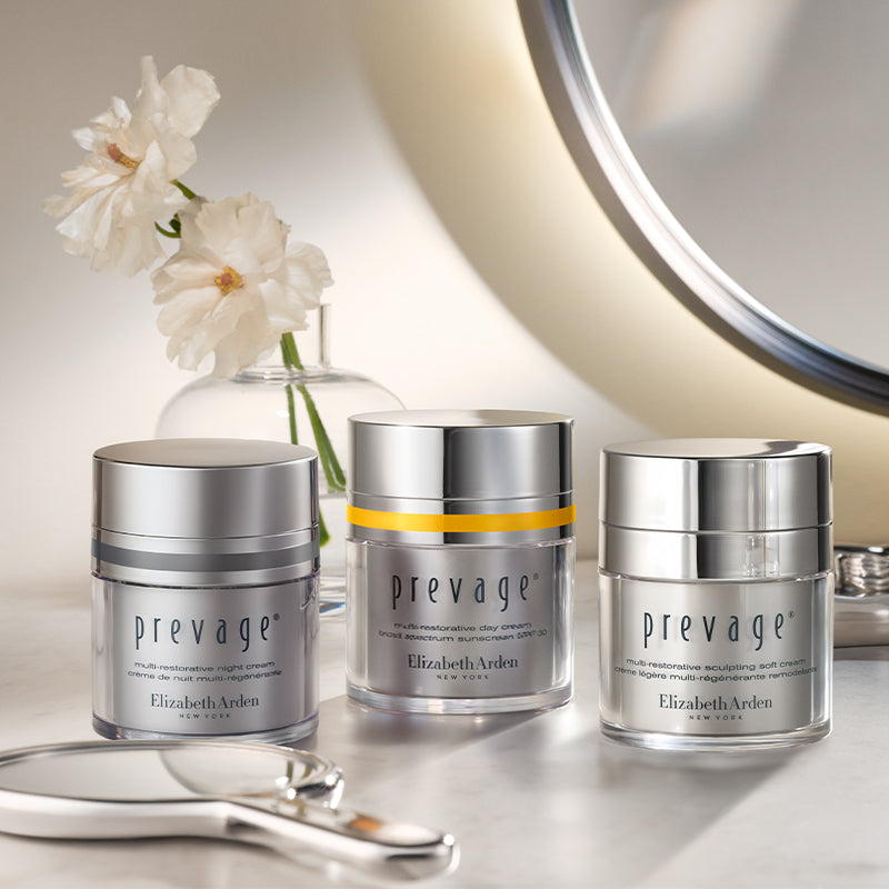 Three Elizabeth Arden Prevage Multi-restorative moisturizers are arranged on a reflective surface with a vase of white flowers and part of a round mirror in the background.