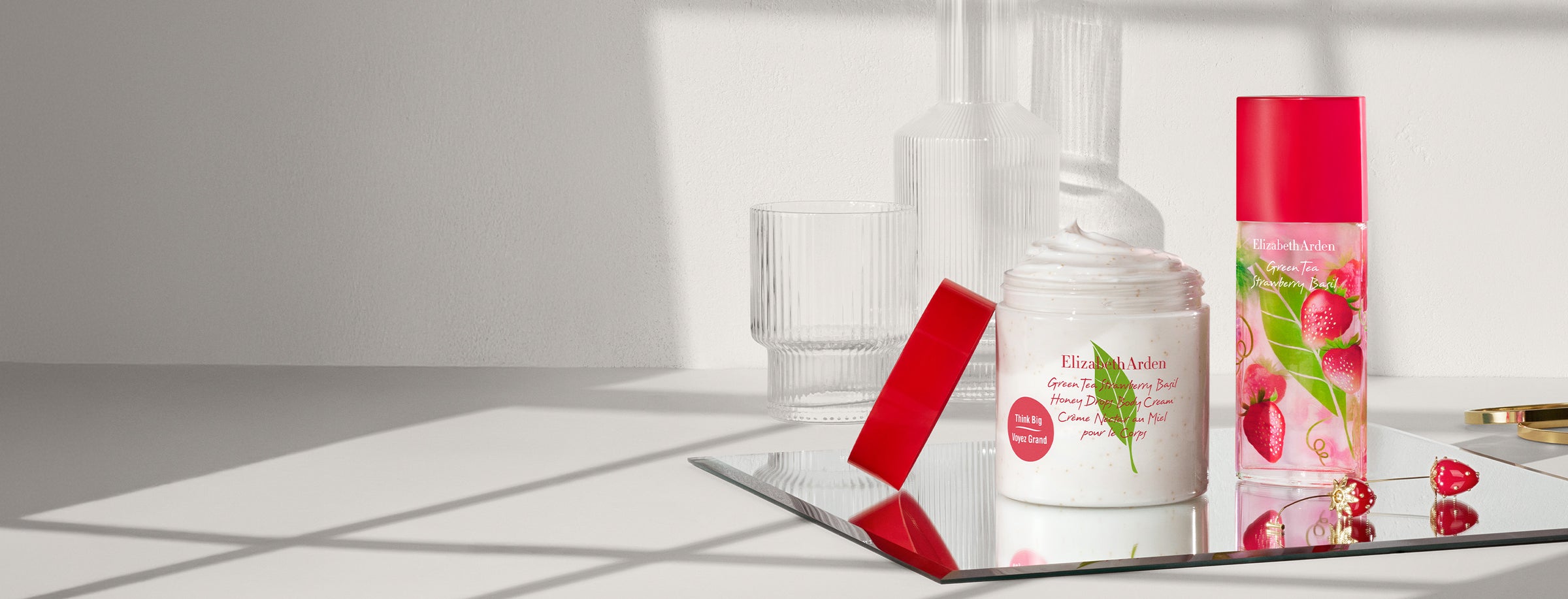 A cosmetic product set on a reflective tray, featuring Elizabeth Arden Strawberry Basil body cream and perfume alongside strawberry earrings and gold bangles.