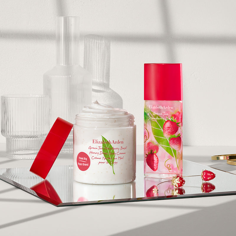 A cosmetic product set on a reflective tray, featuring Elizabeth Arden Strawberry Basil body cream and perfume alongside strawberry earrings and gold bangles.