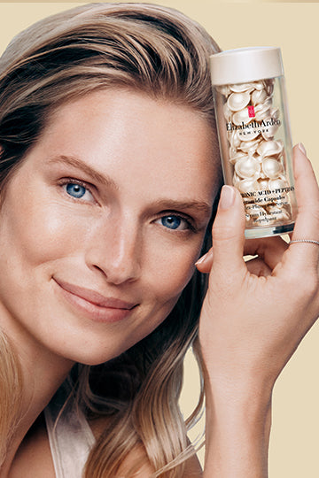 A smiling woman holding Elizabeth Arden Hyaluronic Acid + Peptides Ceramide Capsules Serum on a gold background