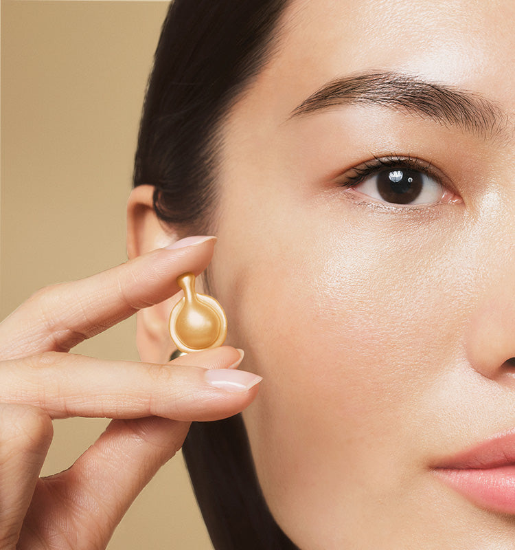 A close-up lifestyle shot of a model  holding a single Advanced Gold Ceramide Capsule close to her ear between two fingers, on a gold background.