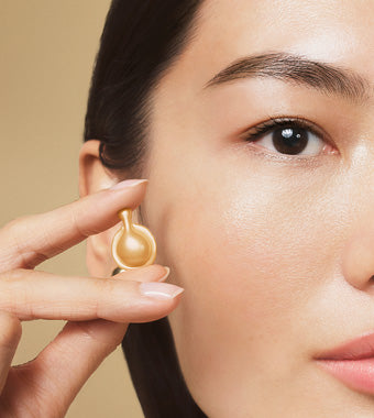 A close-up lifestyle shot of a model  holding a single Advanced Gold Ceramide Capsule close to her ear between two fingers, on a gold background.