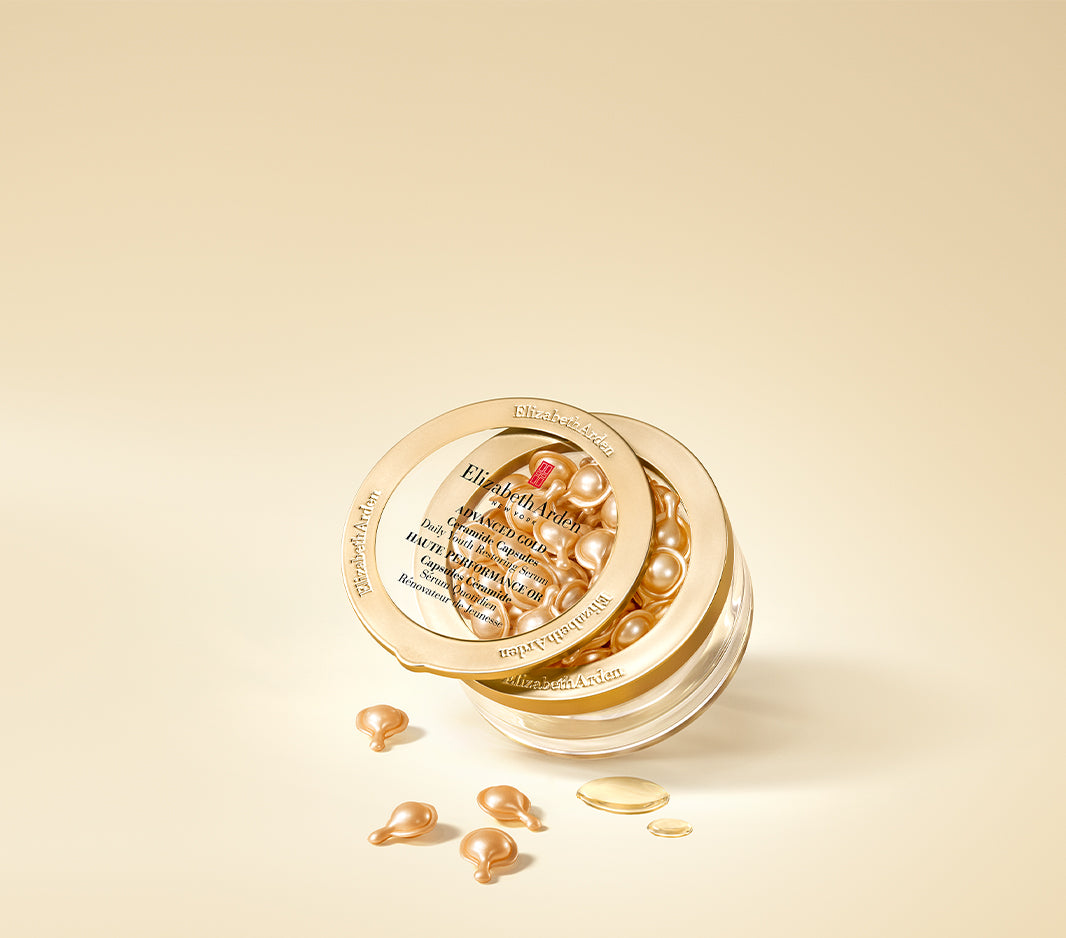 Elizabeth Arden Advanced Gold Ceramide Capsules Daily Youth Restoring Serum in a clear jar with a gold lid, shown on a warm beige background with a few capsules and texture spill scattered in front.