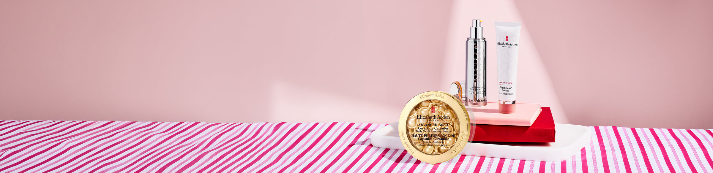 Elizabeth Arden Prevage Anti-aging 2.0 serum, Advanced Gold Ceramide Capsules Serum and Eight Hour Skin Protectant sitting on top of books on a pink background. 