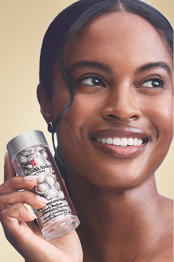 A smiling woman holding Elizabeth Arden Visible Brightening Capsules Serum on a gold background