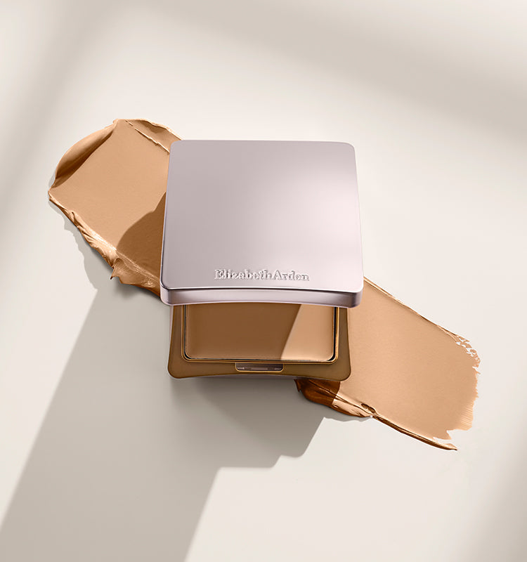 An Elizabeth Arman powder compact in a metallic champagne-gold case, shown open with the pressed powder visible and beige foundation swatches spread on either side on a warm off-white background.