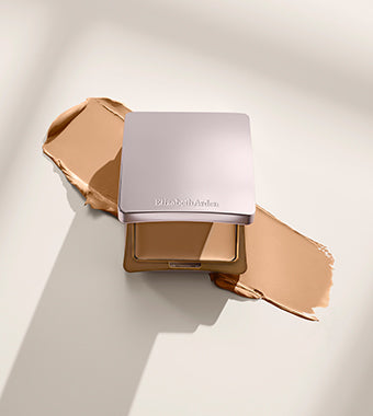 A square makeup compact with a metallic rose-gold lid sits open on a cream-colored surface, surrounded by a smudge of warm beige foundation. The compact brand name 'Blue Silk Skin' is subtly visible on the lid.