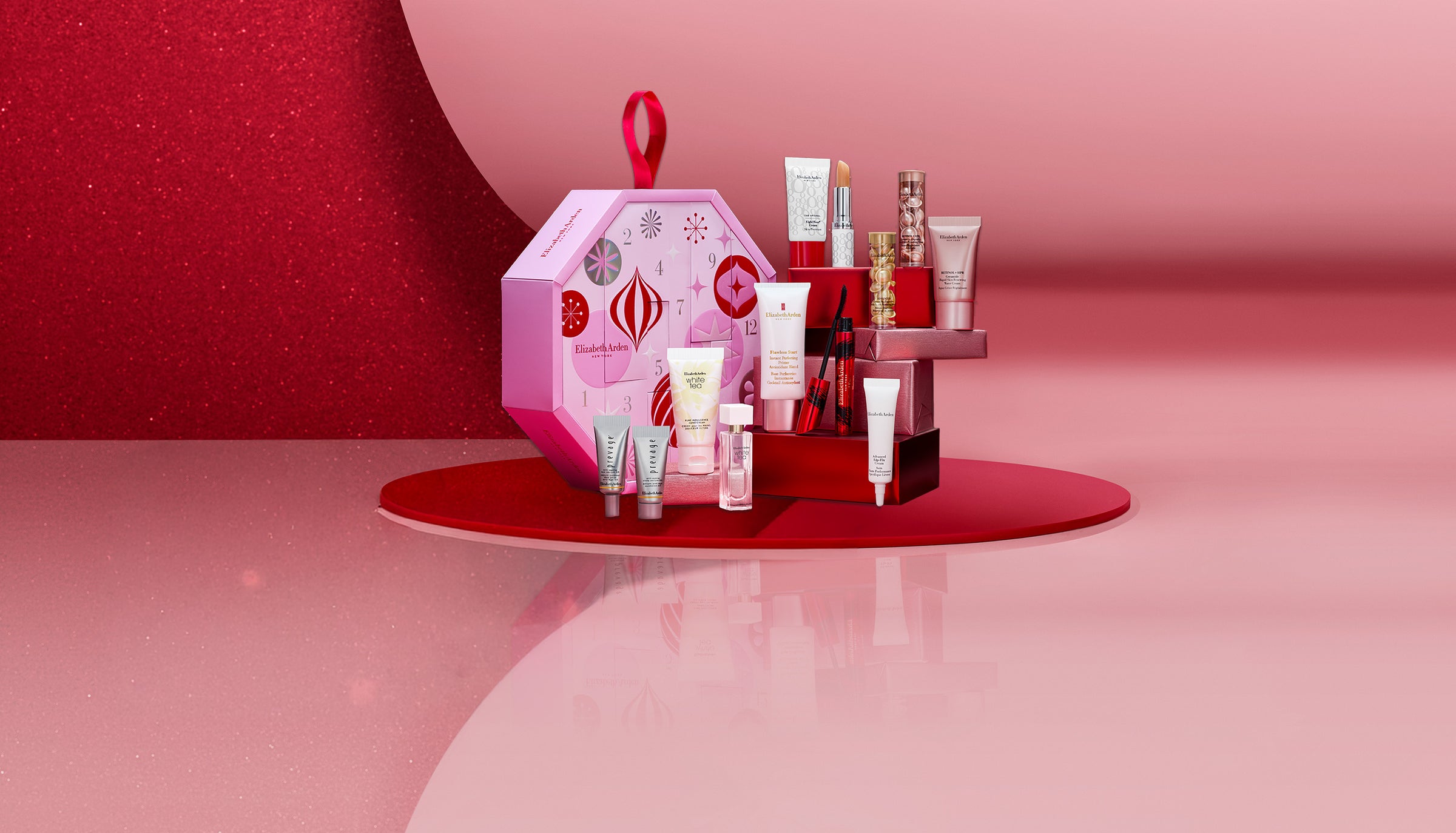 A cosmetics gift set displayed on a red circular pedestal against a pink and red studio background. The set includes a pink patterned gift box with a red loop handle, several red and white tubes, bottles, and jars, and a small red box.