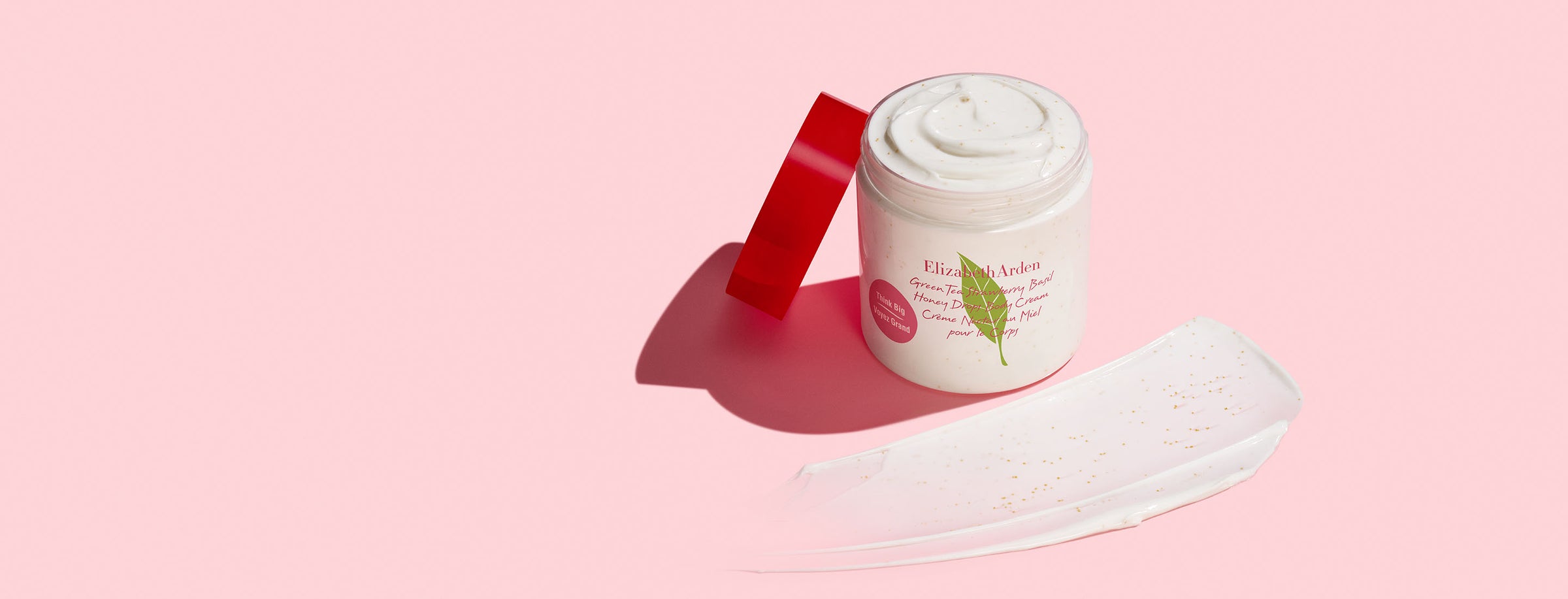 Green Tea Strawberry Basil Honey Drops Body Cream jar with a swatch on a pink background.