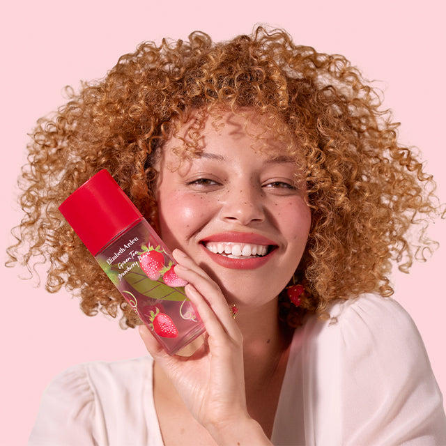 Smiling woman showcasing Green Tea Strawberry Basil Eau de Toilette with a bright pink background.