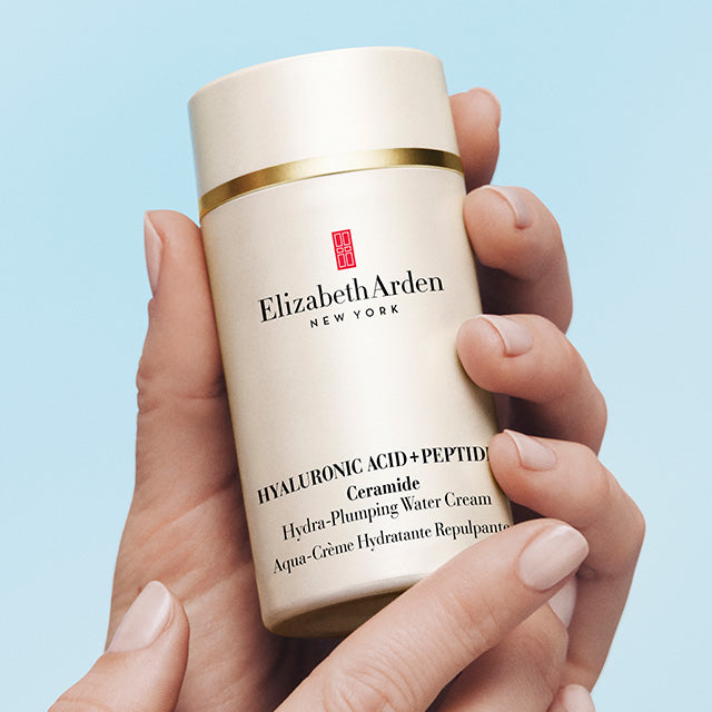 Hand holding a bottle of Elizabeth Arden Hyaluronic Acid + Peptides Water cream against a light blue background